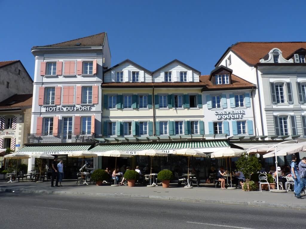 Façade de l'Hôtel du Port et de la Crêperie d'Ouchy sur le quai, volets colorés et terrasses sous stores rayés