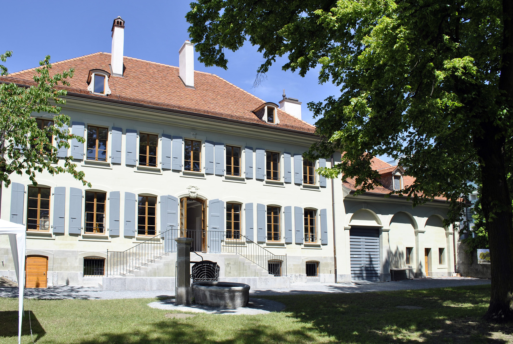 Façade de la Ferme des Tilleuls à volets bleus, avec la fontaine en pierre de la cour