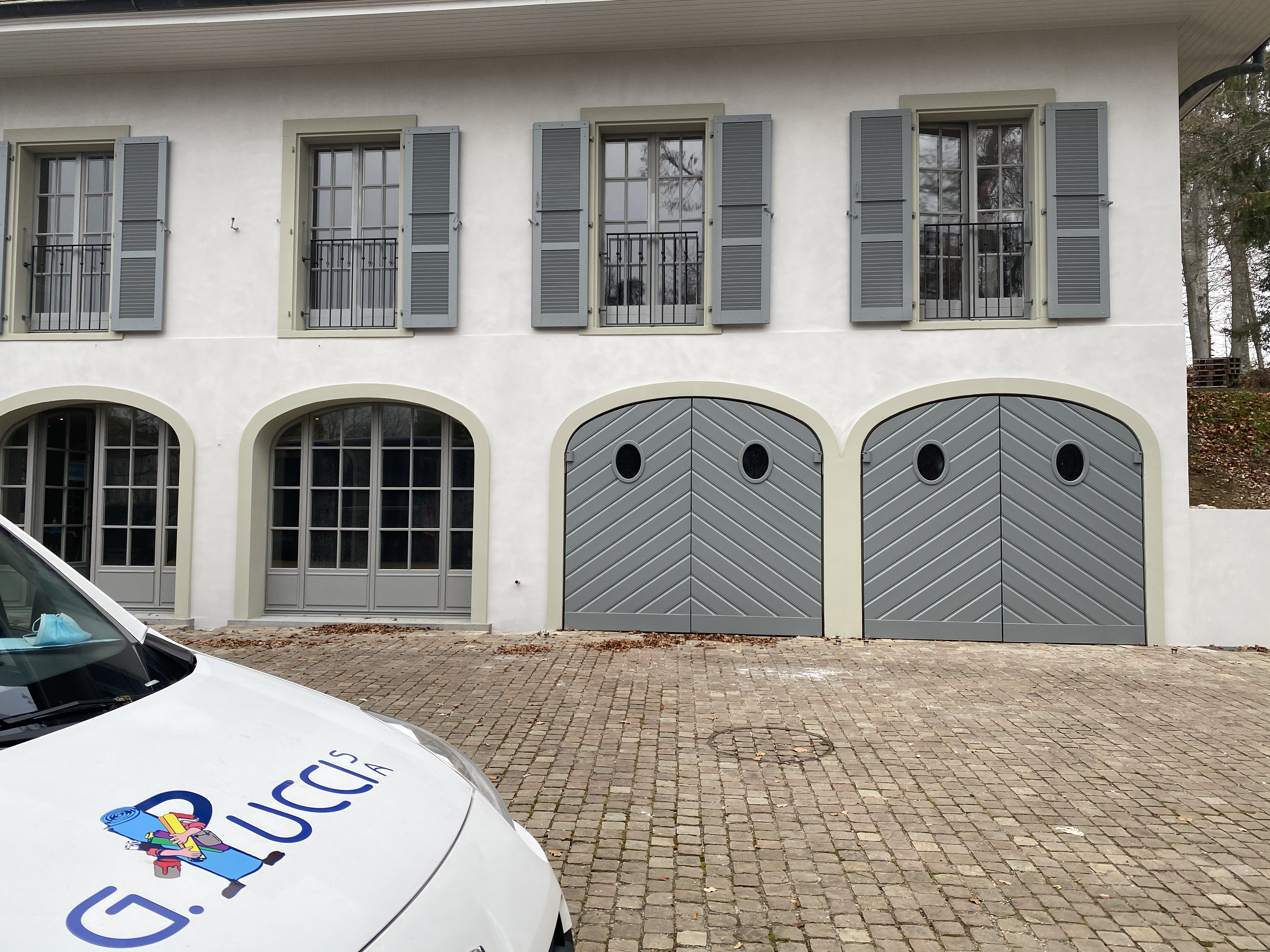 Façade de la maison de maître à volets gris et portes de garage cintrées, avec le véhicule G. Pucci SA