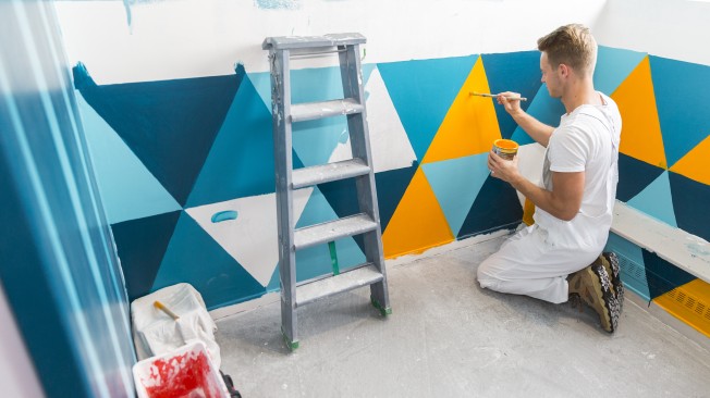 Peintre en tenue blanche appliquant au pinceau un décor géométrique turquoise et jaune sur le mur d'une chambre