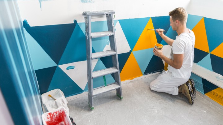 Peintre en tenue blanche appliquant au pinceau un décor géométrique turquoise et jaune sur le mur d'une chambre