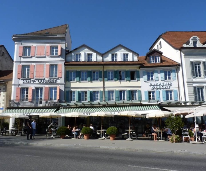 Façade de l'Hôtel du Port et de la Crêperie d'Ouchy sur le quai, volets colorés et terrasses sous stores rayés